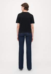 Žena s krátkými kudrnatými vlasy, oblečená v černém svetříku s krátkým rukávem a tmavě modrých bootcut džínách, stojící zády k fotoaparátu na bílém pozadí.