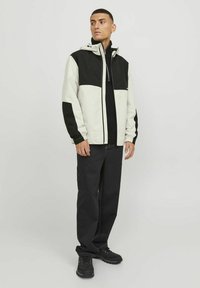 Giacca da uomo a blocchi di colore in crema e nero, materiale impermeabile, chiusura con zip, cappuccio regolabile, con pantaloni neri e calzature texturizzate.