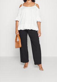 Top bianco off-the-shoulder con orlo arricciato, abbinato a pantaloni neri a gamba larga. La modella tiene una borsa intrecciata color sabbia e indossa tacchi trasparenti.