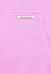 Lyserød sweatshirt med en frontlomme, blød tekstur og et sølvlabel med "bazix republiq." Sømdetaljer langs kanterne.