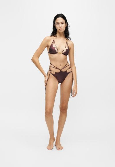 Femme debout pieds nus portant un bikini marron à bretelles fines avec des détails en anneau doré sur un fond clair uni.