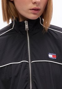 Μαύρο μπουφάν με φερμουάρ, λευκή επένδυση, υφασμάτινη υφή και μικρό κεντητό λογότυπο στο στήθος με την επιγρα φή "TOMMY JEANS".