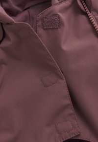 Gros plan sur une veste bordeaux avec une texture lisse, dotée d'une fermeture éclair et d'accents en Velcro. Coutures visibles autour des bords et des points d'attache.