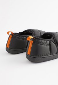 Les chaussons matelassés noirs présentent un accent orange à l'arrière, un design rembourré et une semelle en caoutchouc texturée pour une meilleure adhérence.