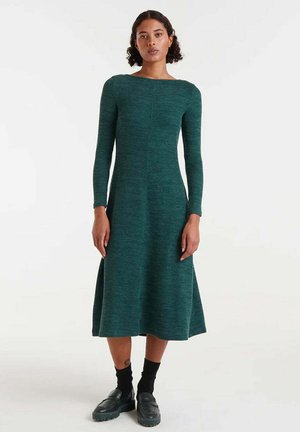 Vestido largo de manga larga en verde oscuro, hecho de una tela de punto texturizada, con escote barco y falda acampanada, combinado con calcetas y zapatos negros.