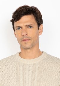 Jasno beżowy sweter dzianinowy z fakturowanym wzorem, z okrągłym dekoltem i subtelnymi wzorami w warkocze, noszony przez mężczyznę z krótkimi ciemnymi włosami.