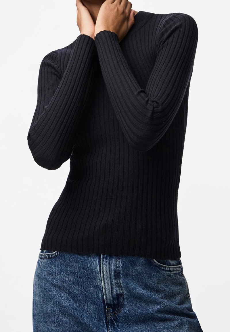 Personne portant un haut noir côtelé à manches longues et un jean bleu, les mains touchant doucement le cou et le visage sur un fond blanc.