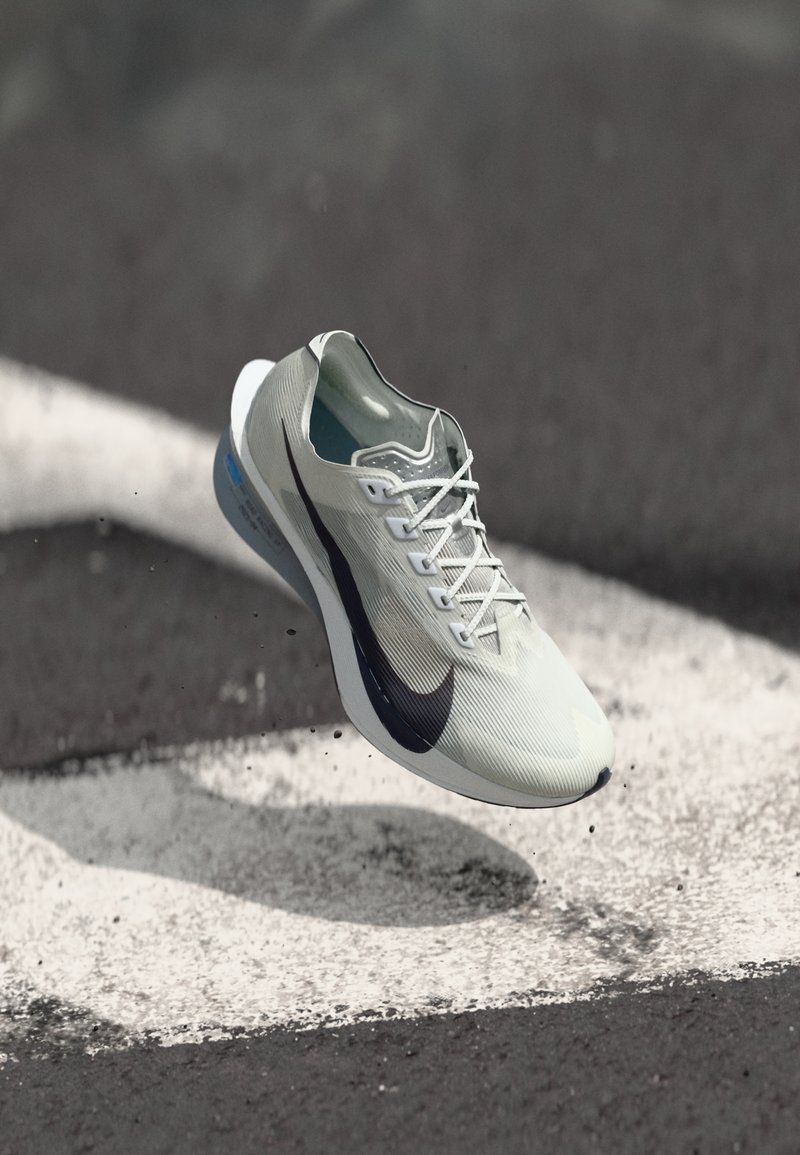 Pantoful de alergare Nike alb și negru, suspendat deasupra unei suprafețe de asfalt cu marcaje rutiere albe și resturi mici.