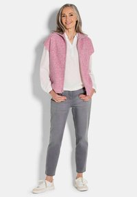 Rosa gestrickte Weste mit Reißverschluss und übergroßen Schultern, kombiniert mit einem weißen Hemd und grauen Slim-Fit-Hosen. Weiße Sneakers runden das Outfit ab.