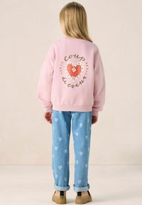 Sweatshirt rose avec un motif en forme de cœur et du texte dans le dos, associé à un jean bleu clair avec un motif de cœurs. Des chaussures beiges complètent la tenue.