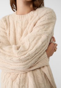 Kermanvärinen palmikkoneulepusero, jossa on pyöreä pääntie, tekstuurikuvioita ja pehmeä, pörröinen materiaali sekä resorit hihansuissa ja helmassa.