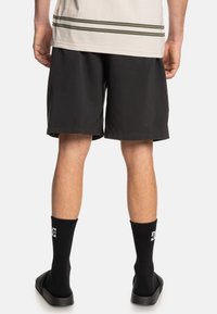 Black shorts made of lightweight fabric, standard fit, with a smooth texture. Worn with black socks featuring a white logo and sandals.