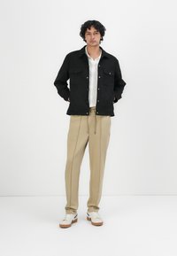 Schwarze Baumwollhemdjacke mit zwei Brusttaschen, getragen über einem weißen strukturierten Hemd, kombiniert mit beigen Hosen und weißen Sneakers mit braunen Akzenten.