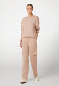 Felpa in pile rosa con logo "Guess", abbinata a pantaloni cargo coordinati con tasche laterali. L'outfit ha una vestibilità rilassata e una texture morbida.