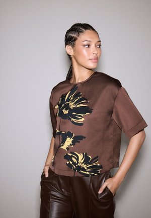 Top marrone con motivo floreale, maniche corte; presenta disegni neri e gialli, una scollatura rotonda e una vestibilità comoda, abbinato a pantaloni marroni.