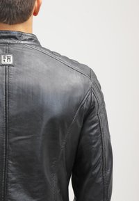 Chaqueta de cuero negro con una textura suave, costuras verticales y una etiqueta cuadrada en la parte posterior con iniciales, diseñada con una forma moderna.
