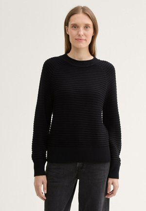 Black ribbed knit sweater with long sleeves, round neckline, and textured pattern. Paired with dark jeans, has a fitted silhouette.