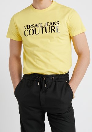 Mężczyzna w żółtej koszulce z napisem "Versace Jeans Couture" i czarnych spodniach z trokami, z rękami w kieszeniach, na neutralnym tle.