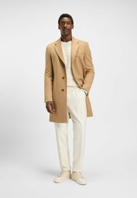 Brun uldfrakke med dobbeltknapdesign, beige sweater under, parret med hvide bukser og lyse sneakers. Ensfarvet baggrund.