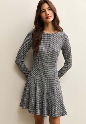 Robe pied-de-poule en noir et blanc avec manches longues, corsage ajusté, jupe évasée et encolure ronde. Confectionnée dans un tissu texturé.