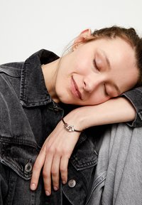 Bracelet en cuir marron avec fermoir en argent présentant un design de boucle entrelacée. Le modèle porte une veste en denim gris, reposant sa tête sur son bras.