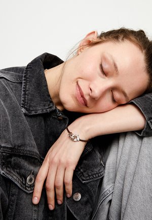 Bracelet en cuir marron avec fermoir en argent présentant un design de boucle entrelacée. Le modèle porte une veste en denim gris, reposant sa tête sur son bras.