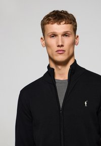 Zwarte zip-up sweater met een hoge kraag, gemaakt van gebreid materiaal. Heeft een geborduurd logo op de linkerborst. Draagt over een grijze shirt.