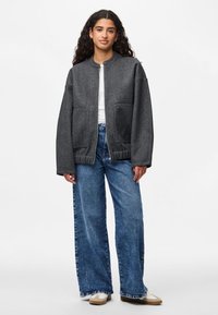 Giacca bomber grigia con colletto rotondo, due tasche frontali e polsini a coste, abbinata a jeans blu a gamba larga e sneakers beige.