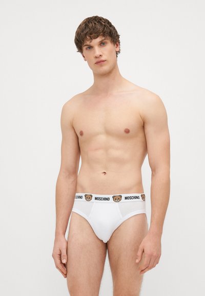 Bela moška spodnja perila z črnim pasom, na katerem je napis "MOSCHINO" in logotip medveda. Tkanina se zdi gladka, z oprijetim fitom okoli pasu.