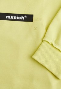 Tessuto di felpa a coste di un giallo chiaro, con un'etichetta nera su cui è riportato il testo "mxnich". La manica presenta un dettaglio con bordo sfilacciato.