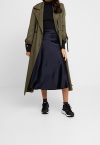 Zielona oliwkowa trench coat, prążkowany czarny top, granatowa satynowa spódnica, czarne buty sportowe. Cechy to długie rękawy i rozkloszowana spódnica.