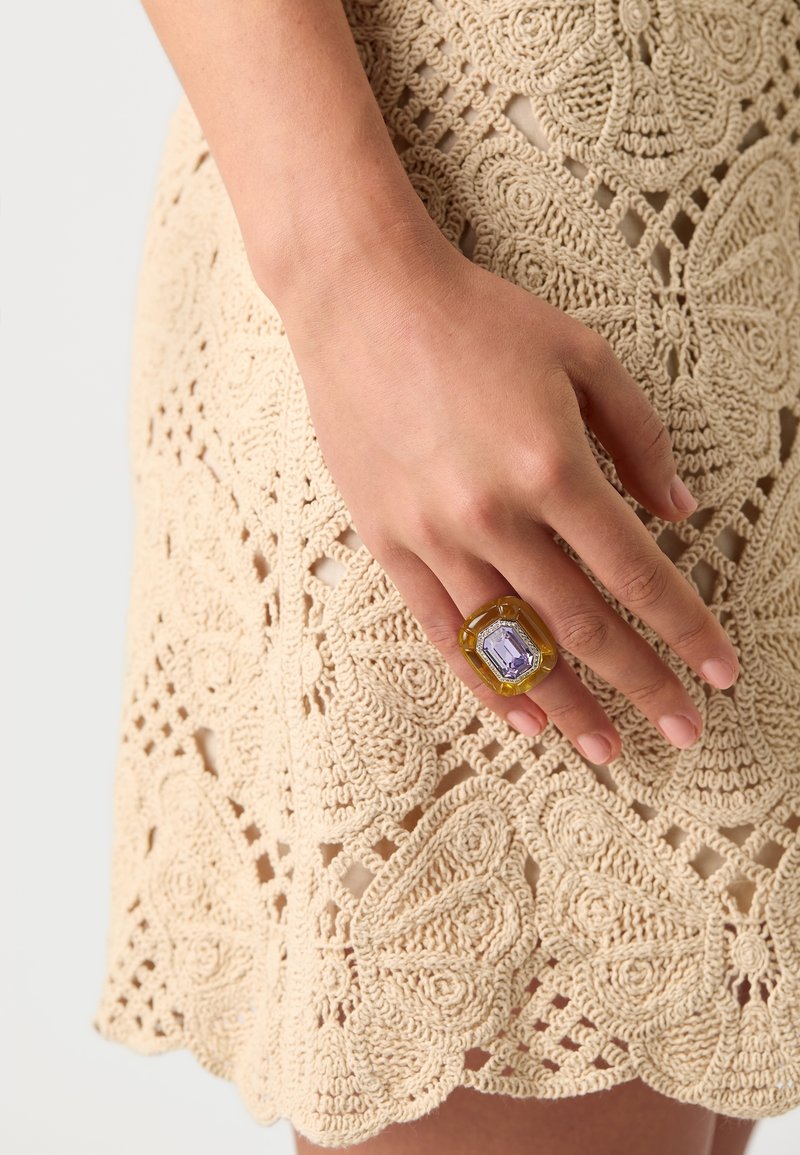 Main reposant sur une robe en crochet beige, portant une grande bague rectangulaire avec une pierre centrale violette et une bordure jaune.