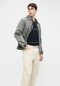 Νεαρός άνδρας που φοράει γκρι τζιν μπουφάν πάνω από μαύρο πουκάμισο και μπεζ παντελόνι, με το ένα χέρι στην τσέπη, κοιτάζοντας προς το πλάι.