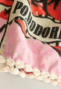 Tissu en coton avec un fond rose présentant un motif de tomate et des lettres noires. Fini avec un bord en crochet blanc.