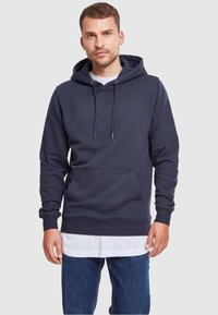 Laivastonsininen huppari pehmeästä, teksturoidusta materiaalista, jossa on kengurutasku, kiristysnauhallinen huppu, sekä resorit helmassa ja hihoissa, kerrospukeutuneena valkoisen paidan päälle.