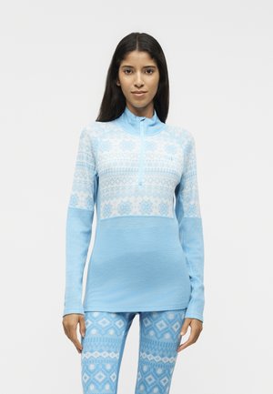 Mujer de pie vistiendo un top con cremallera de manga larga con estampado azul claro y leggings a juego sobre un fondo blanco liso.
