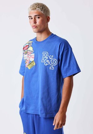 Jeune homme aux cheveux blonds courts portant un t-shirt bleu et un pantalon, le t-shirt présente des patchs graphiques colorés et de grandes lettres blanches "Pxp".