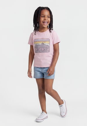 Niña sonriente con cabello trenzado, lleva una camiseta rosa de Patagonia, pantalones cortos de mezclilla y zapatillas blancas, caminando frente a un fondo sencillo.