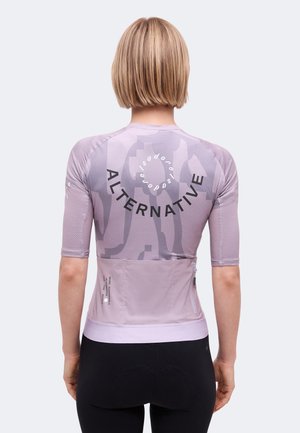 Person mit kurzen blonden Haaren, die ein eng anliegendes lavendelfarbenes Fahrradtrikot mit dem Schriftzug „ALTERNATIVE“ und Rückentaschen trägt, kombiniert mit schwarzen Radhosen.
