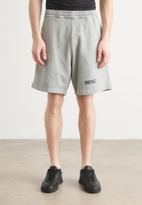 Pantalones cortos de algodón gris con cintura elástica, bolsillos laterales y un logo negro en el dobladillo. Llevados con zapatillas negras sobre un fondo neutro.