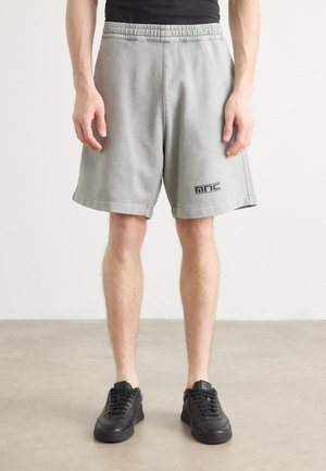 Gray cotton shorts with an elastic waistband, side pockets, and a black logo on the hem. Worn with black sneakers on a neutral background.