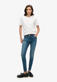 T-shirt blanc à manches courtes associé à un jean skinny bleu, avec une ceinture noire et des mocassins noirs. Textures lisses de coton et de denim.