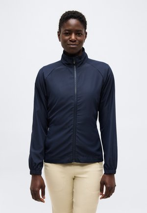 Woman wearing a navy blue zip-up jacket with high collar and beige pants, standing against a plain white background.