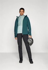 Kurtka zapinana na zamek w kolorze morskim i jasnoniebieskim, noszona na lekkim topie, w połączeniu z czarnymi spodniami i czarnymi butami; trzyma szary kask.