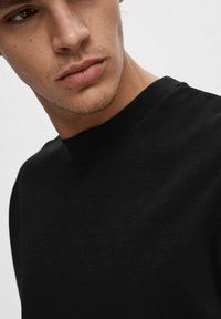 Tricou negru din bumbac, cu guler rotund, având o textură netedă, un design simplu și o siluetă ajustată. Vedere detaliată a gulerului și a părții superioare a țesăturii.