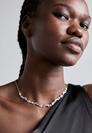 Collier en argent avec un design floral, orné de petites pierres précieuses transparentes, tombant doucement autour du cou sur un fond de tissu sombre.