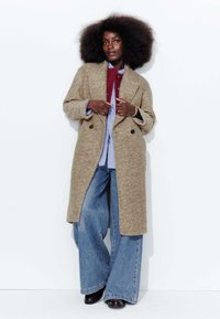 Manteau en laine à motif pied-de-poule beige avec un col à revers, associé à une chemise bleue et un jean en denim à jambe large. Le mannequin se tient devant un fond blanc.