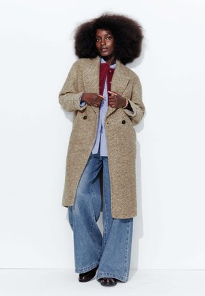 Manteau en laine à motif pied-de-poule beige avec un col à revers, associé à une chemise bleue et un jean en denim à jambe large. Le mannequin se tient devant un fond blanc.