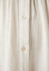 Ανοιχτό μπεζ υφασμάτινη επιφάνεια με υφή και υφαντό σχέδιο, διαθέτει κατακόρυφη κουμπότρυπα και δύο στρογγυλά κουμπιά με τέσσερις τρύπες.