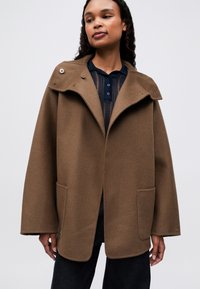 Mujer vistiendo un abrigo marrón holgado sobre una blusa de encaje azul marino y pantalones oscuros, de pie contra un fondo claro y liso.